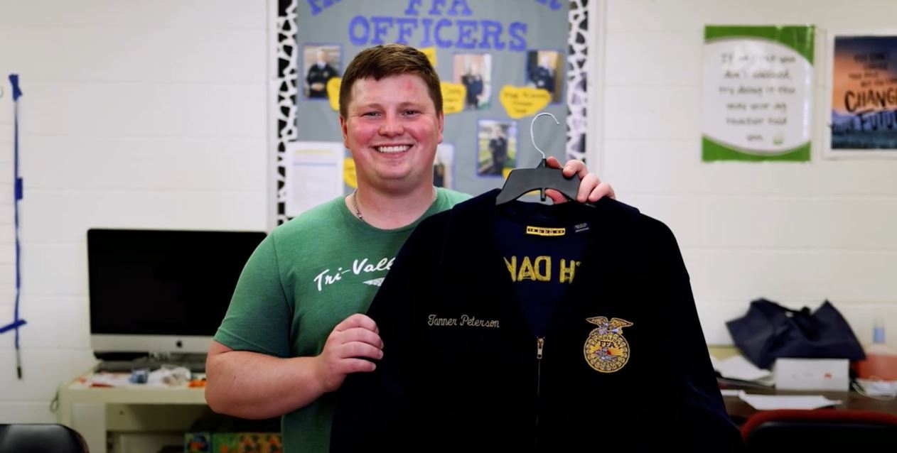 A smiling young man holding up a hanger with a navy blue jacket with a FFA patch on it